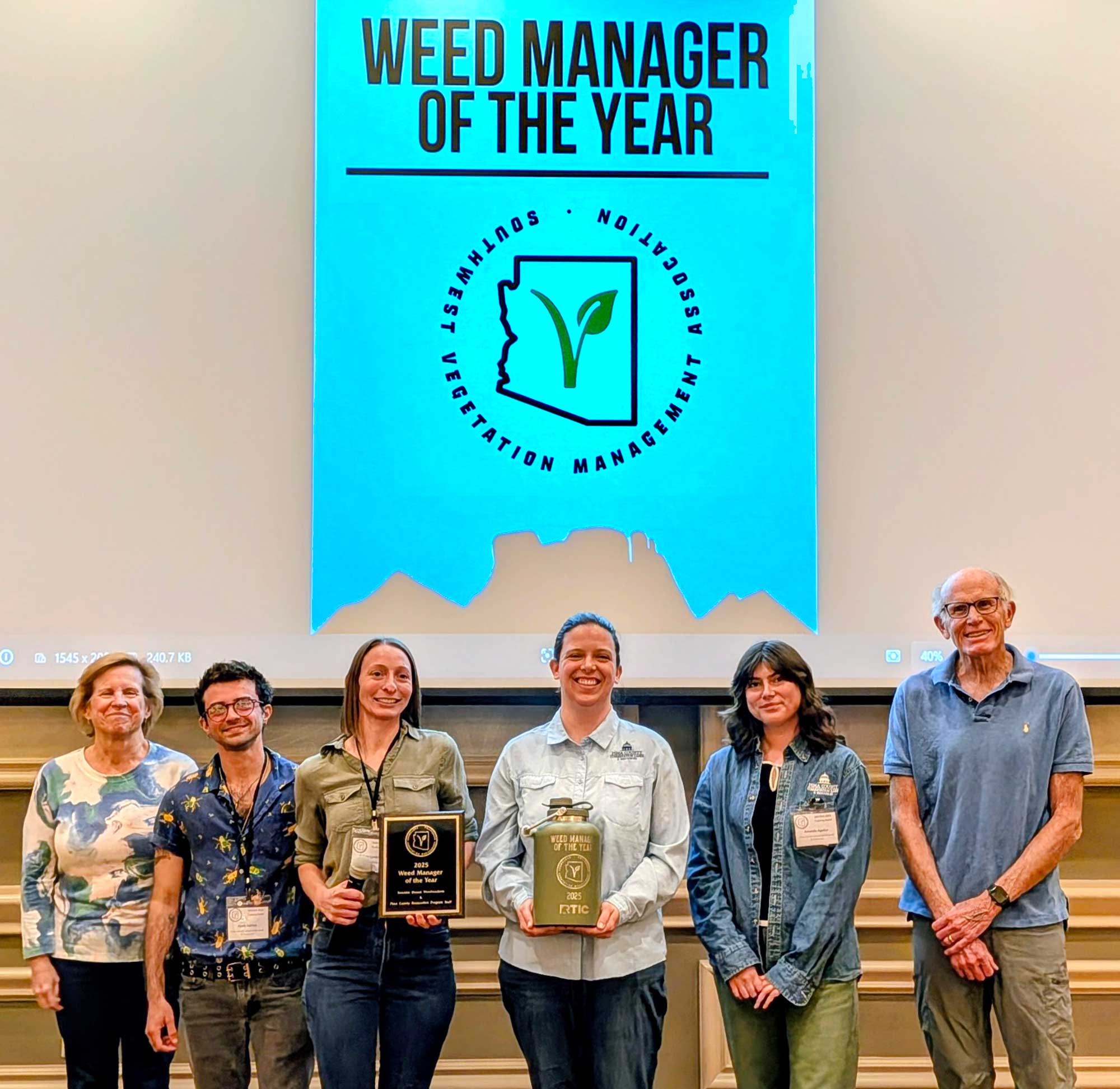 six people holding two plaques standing under a Weed Manager of the Year banner